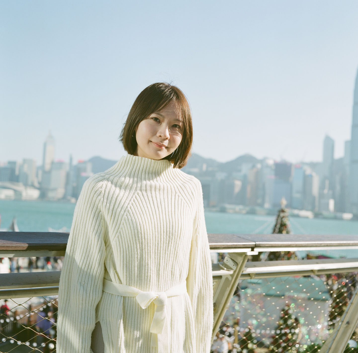 Young woman in cozy white sweater posing with Victoria Harbour skyline in background, sunlight and Christmas decorations at WKCD, Hong Kong.