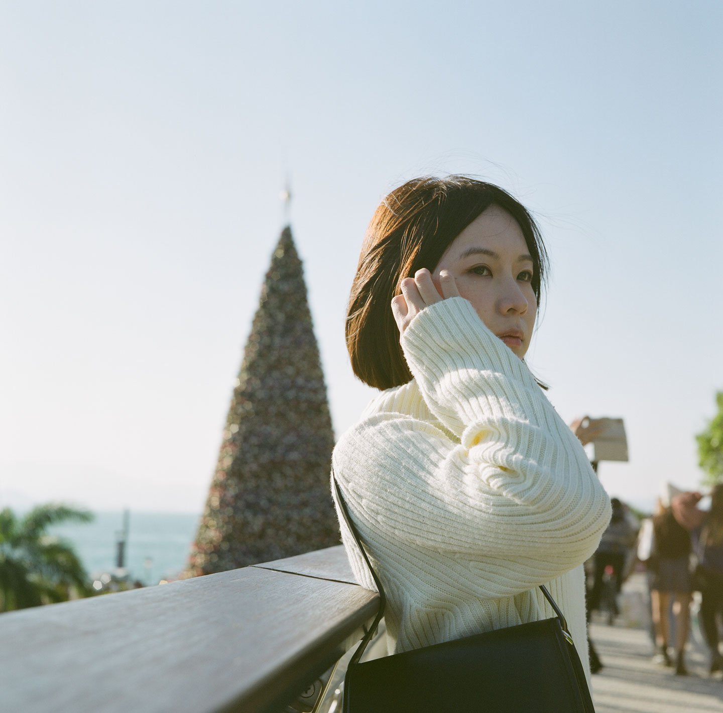 Young woman turning towards camera in white sweater, Christmas tree and winter sunlight at WKCD, Hong Kong, city park in background.