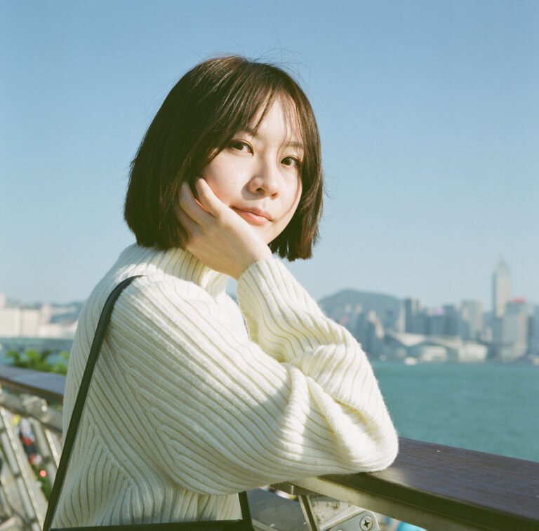 Young woman with short hair in a white sweater resting on railing, Victoria Harbour skyline and blue sky in background, outdoor portrait at WKCD, Hong Kong.