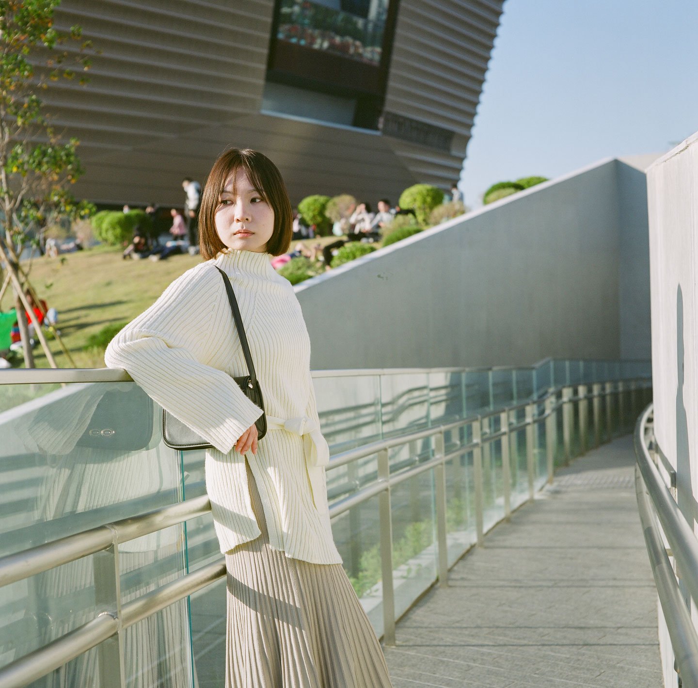 Young woman in a white sweater posing by glass railings at WKCD, Hong Kong, with modern architecture, outdoor garden, and natural sunlight.