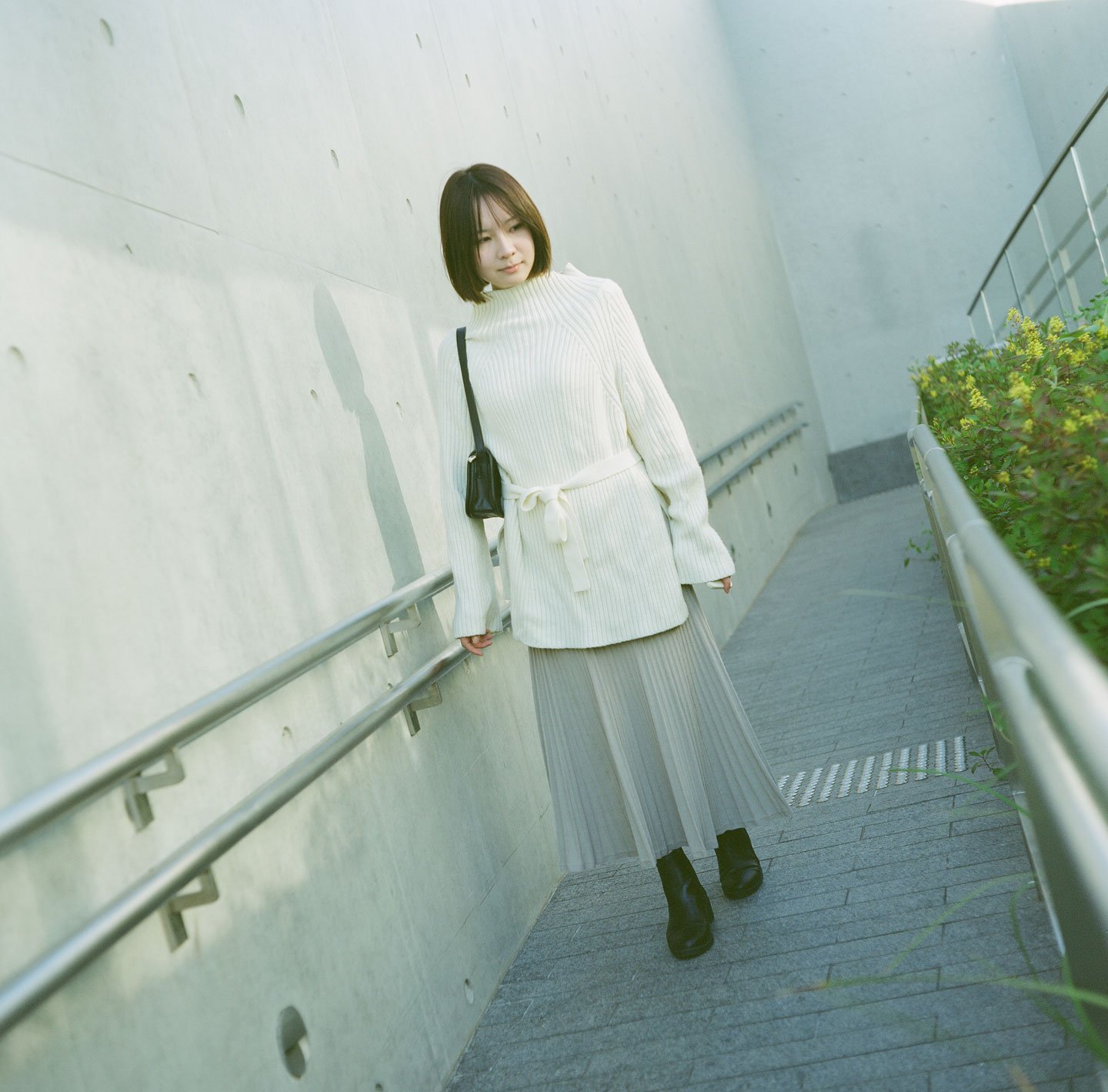 Young woman in a white sweater posing outdoors at WKCD, Hong Kong, with soft sunlight, grey wall, and winter fashion style.