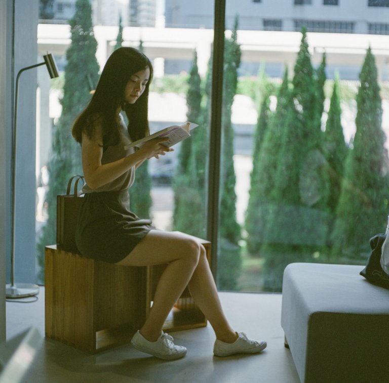 Woman sitting indoors by a window, reading a book, with tall green trees and cityscape in the background at Oi! Art Space, Hong Kong—part of Urban Elegance series (12/13).