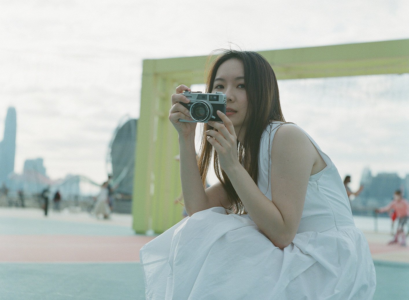 Woman in white dress holding a film camera at East Coast Park Hong Kong with cityscape background